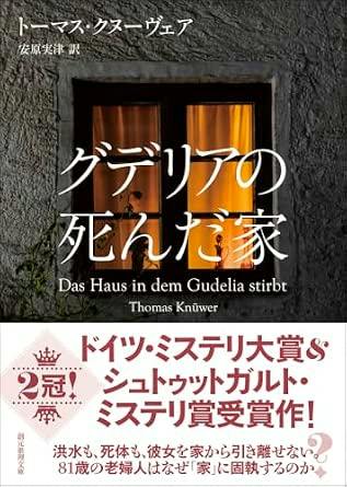 Front cover of the book showing a lit window set in a textured stone wall with the Japanese title 'グデリアの死んだ家' overlaid and a promotional white band at the bottom.