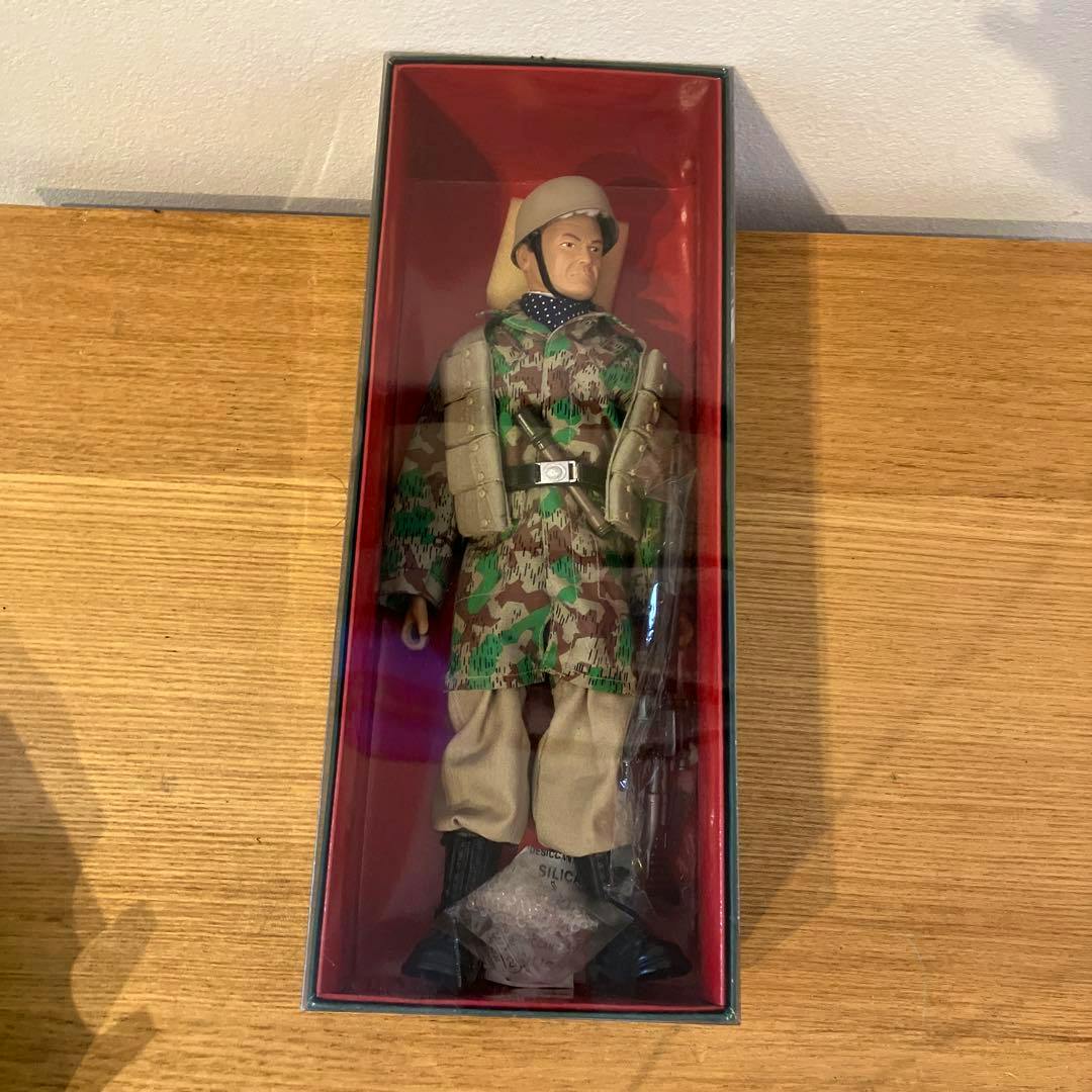 Boxed 1/6 scale military action figure wearing a camouflage jacket and tan trousers visible through the display window.