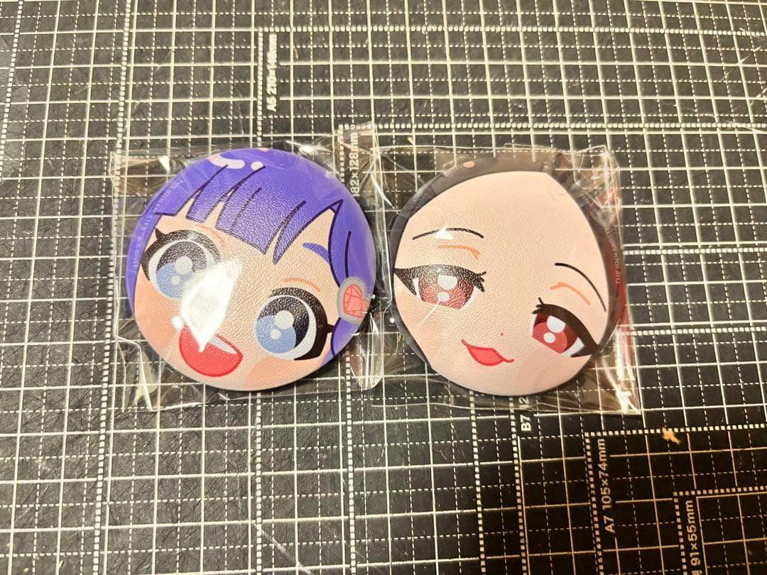 Two round 'ぷにぷに' can badges showing printed anime faces in plastic wrappers on a gridded cutting mat.