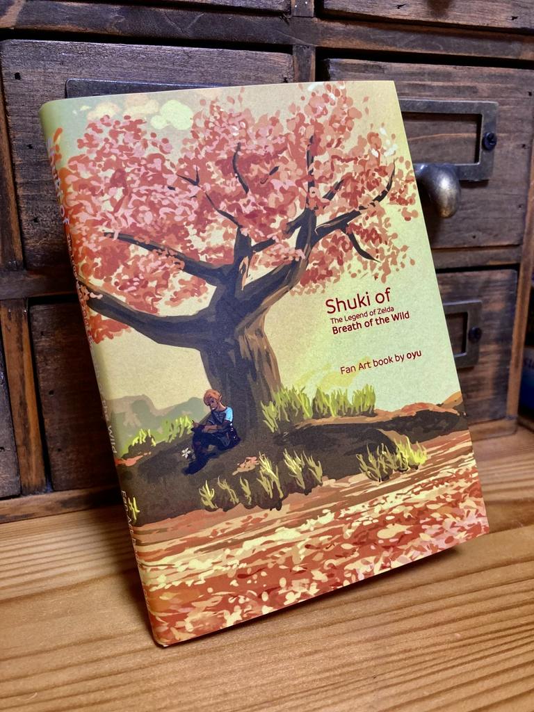 Photograph of the illustration book front cover titled “Shuki of Breath of the Wild,” showing a tree with pink leaves and a character sitting beneath it.