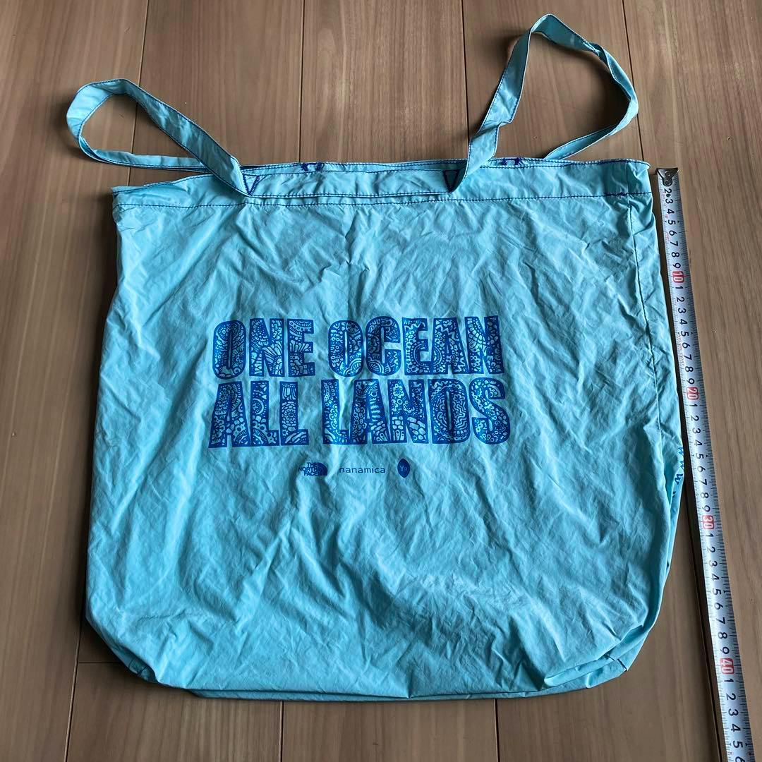 Light blue reusable tote bag laid flat showing the "ONE OCEAN ALL LANDS" print with a measuring tape beside it on a wooden floor.