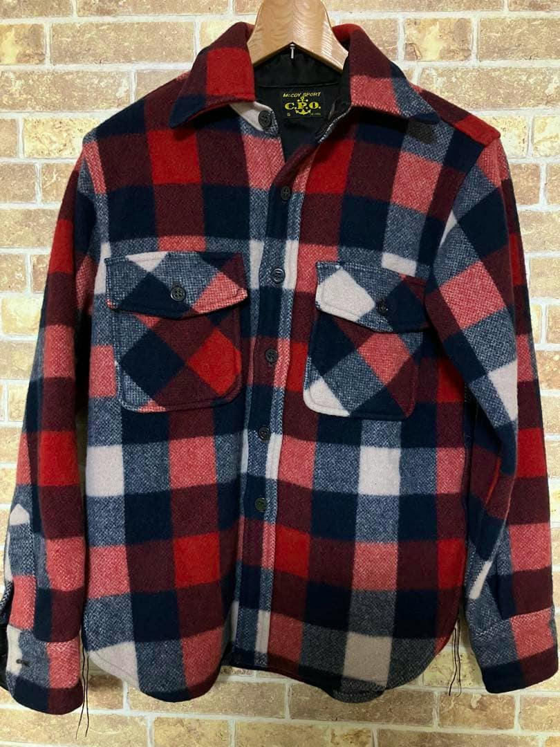 Front view of a red, navy, and white checkered button-up shirt jacket on a wooden hanger against a brick wall.