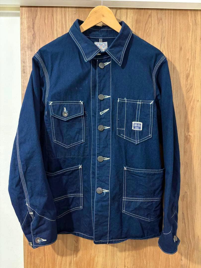Front view of a dark blue coverall-style men's shirt with white contrast stitching, multiple patch pockets, and buttons hanging on a wooden hanger.