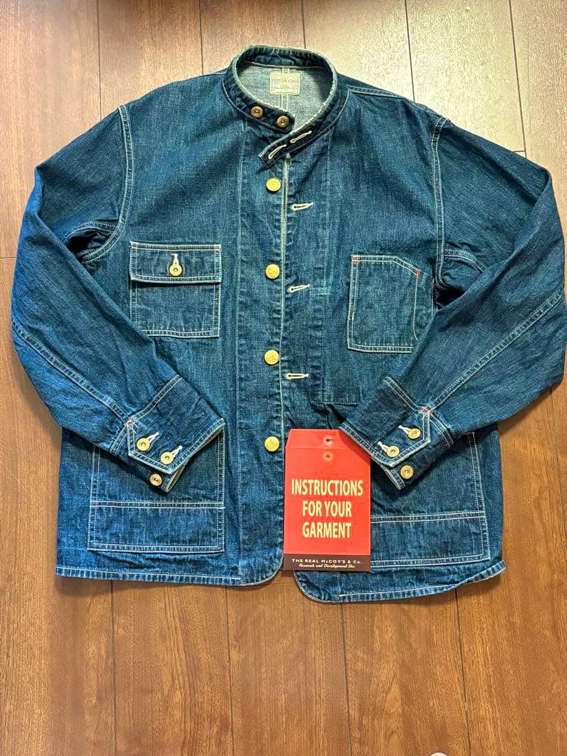 Front view of a blue denim coverall jacket laid flat on a wooden floor with an instruction tag visible near the hem.