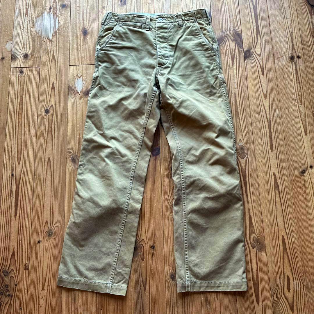 Beige cotton men's chino pants laid flat showing the front view on a wooden floor.