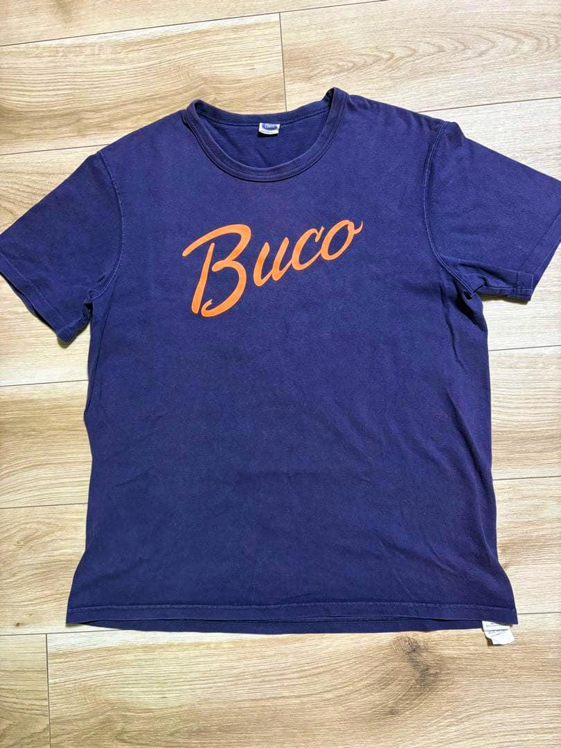 Navy T-shirt laid flat on a wooden floor with an orange "Buco" script printed on the front.