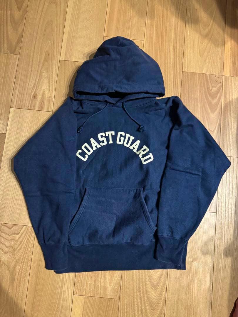 Navy cotton hoodie with "COAST GUARD" printed on the chest laid flat on a wooden floor.