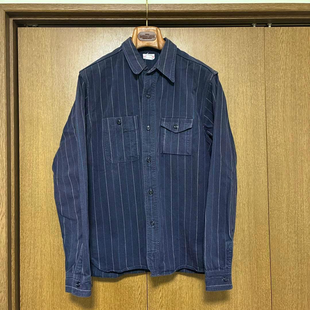 Front view of a navy blue pinstriped men's cotton work shirt (The REAL McCOY'S) size 15 hanging on a wooden hanger against wooden doors.
