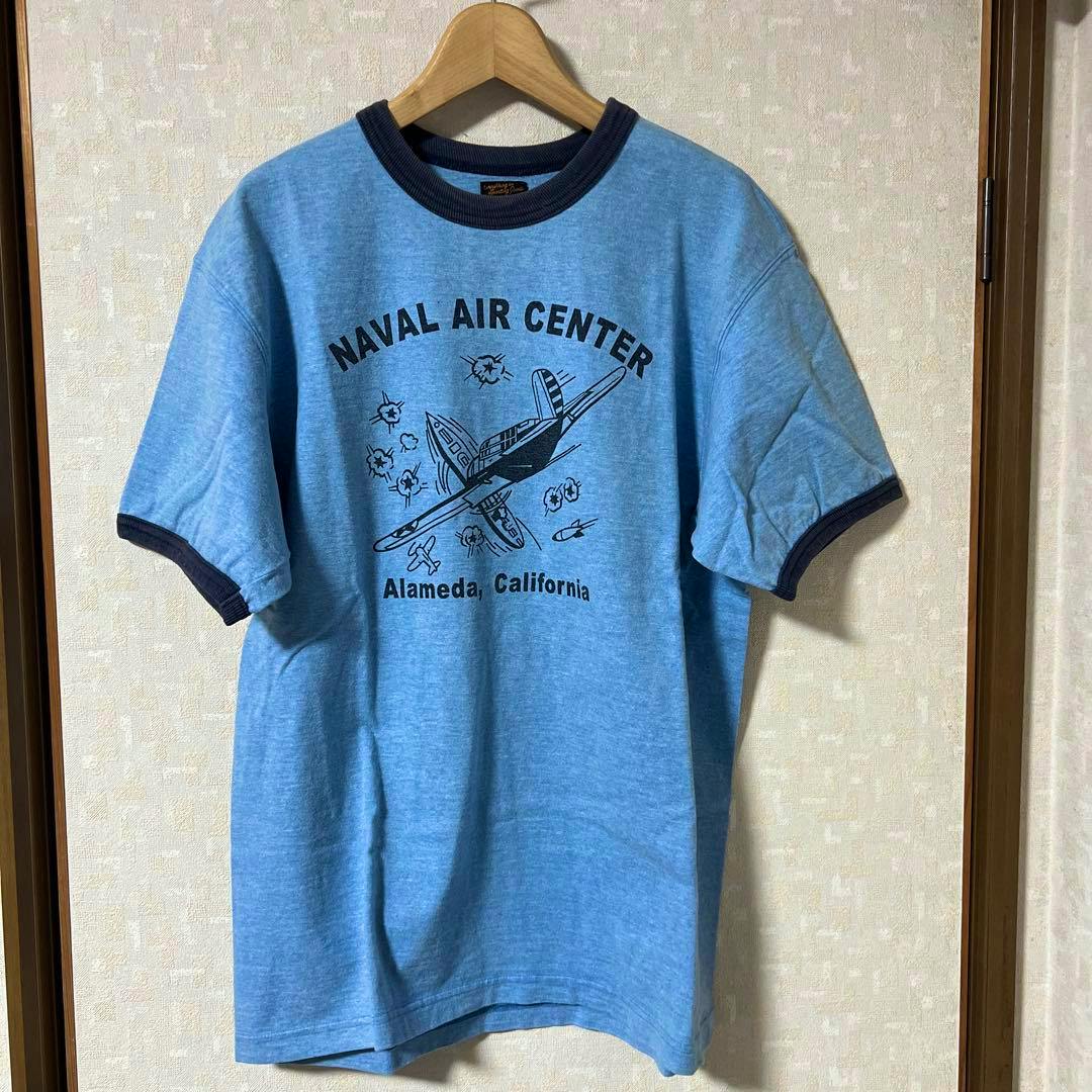 Blue men's T-shirt with navy collar and "Naval Air Center" airplane graphic hanging on a wooden hanger.
