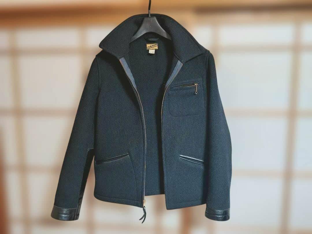 Front view of a men's jacket hanging on a hanger showing a center zipper, a zippered chest pocket, and two lower pockets.