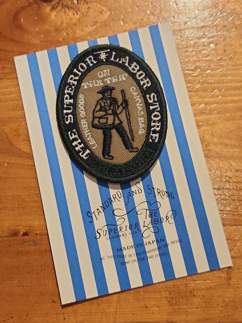 Oval embroidered patch labeled "The Superior Labor Store" displayed on a blue-and-white striped card atop a wooden surface.