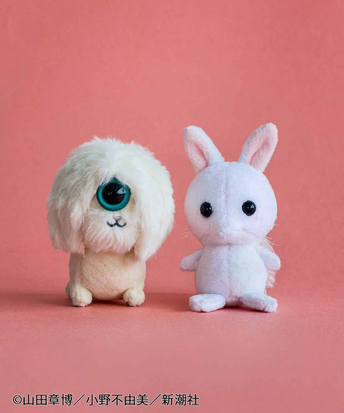 Photo of the 使令マスコット〈什鈷・雀胡〉 plush mascots showing two small plush figures on a pink background: a beige long‑haired figure with one large green eye and a white rabbit‑like figure with black eyes.