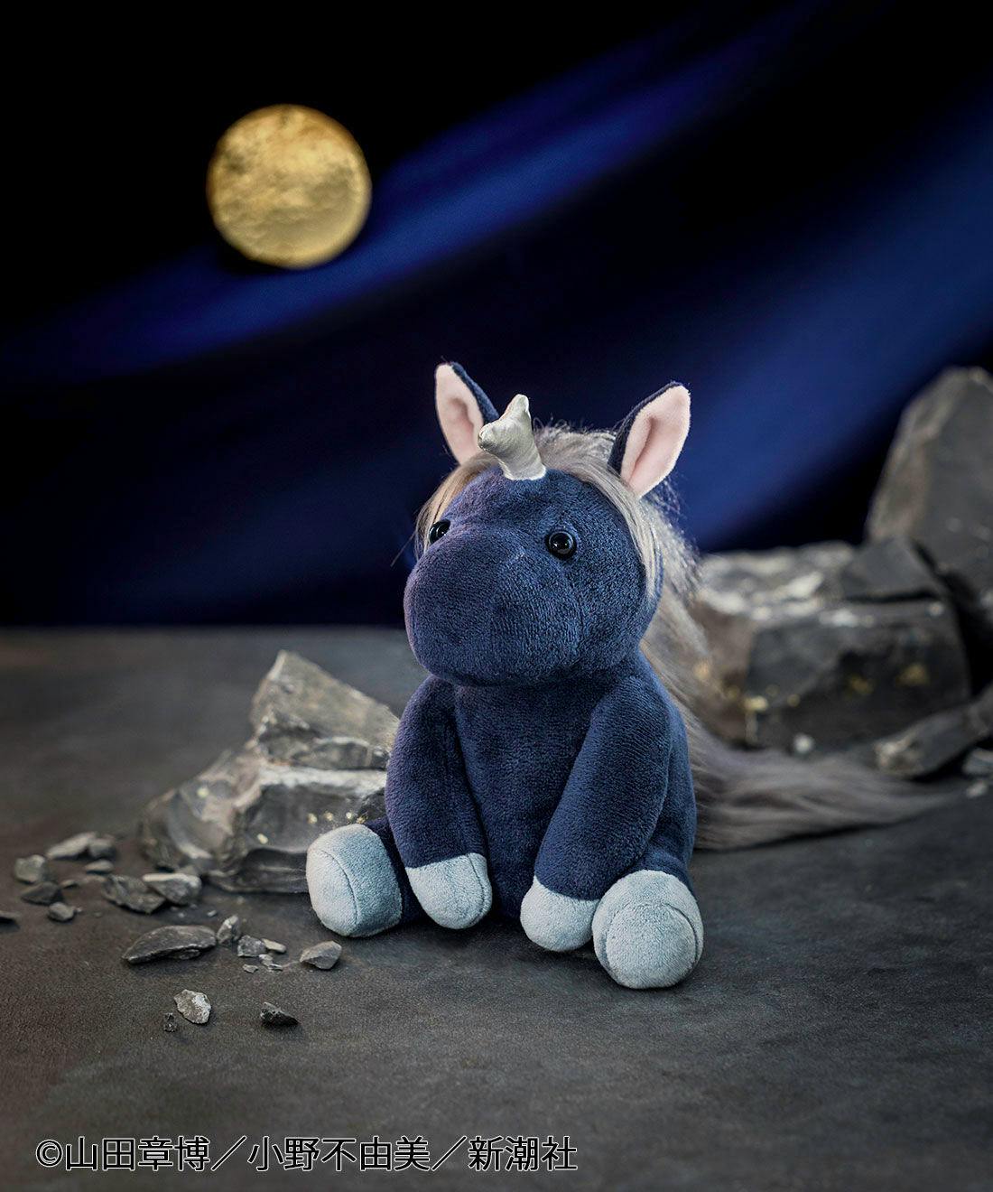 A navy-blue 泰麒 plush toy with a small horn, pale hooves, and a mane seated among rocks with a moon backdrop.