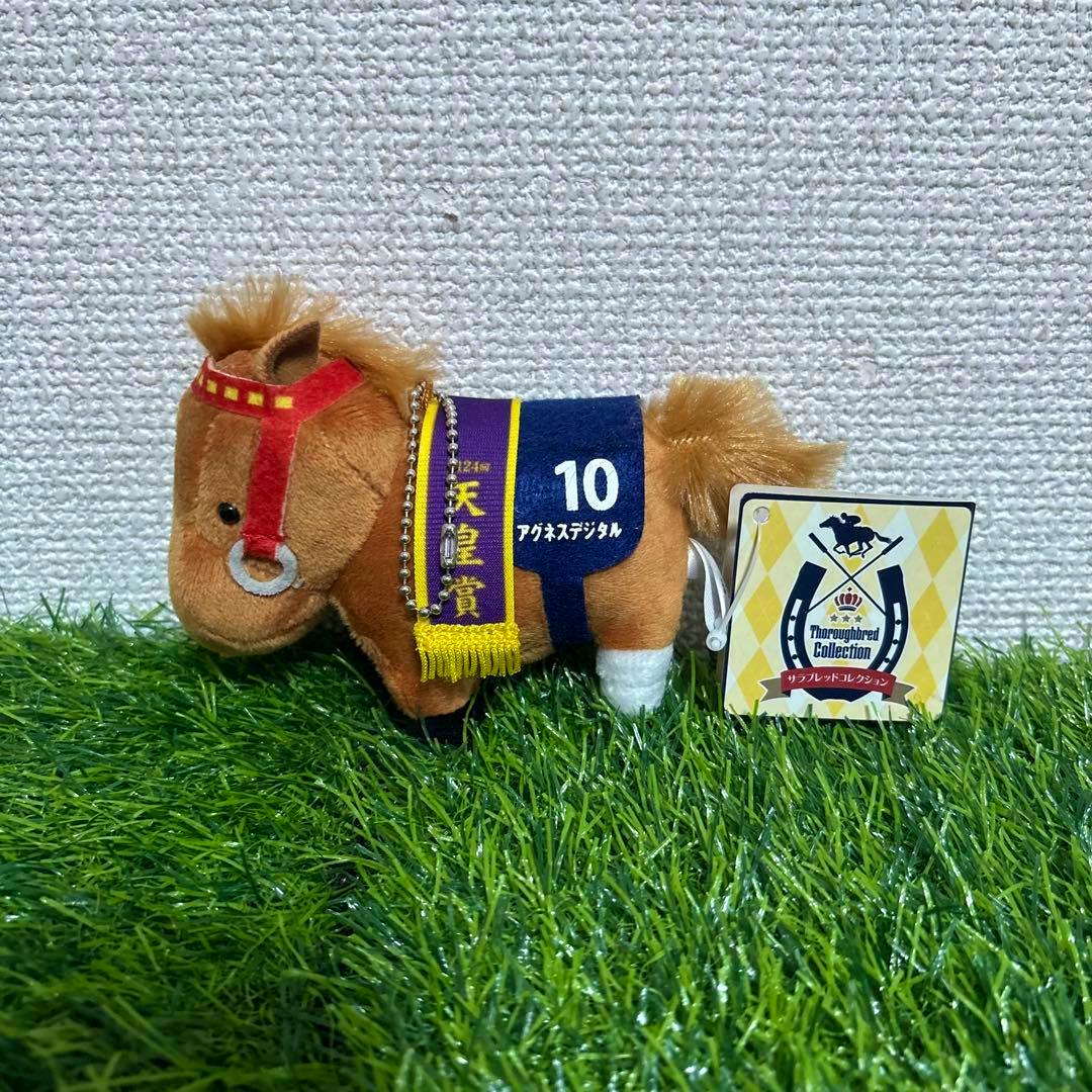 Plush horse mascot keychain with a blue saddle marked “10”, purple sash and attached tag, shown on green artificial grass.