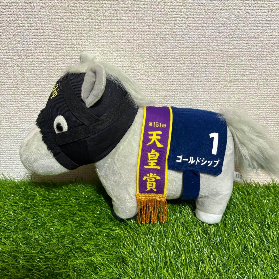 Side view of a Gold Ship plush toy wearing a black hood, a purple sash, and a blue number "1" cloth on artificial grass.