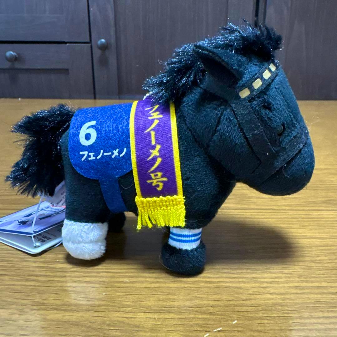 A plush mascot ball-chain horse labeled "フェノーメノ" shown from the side with a blue saddle cloth numbered 6 and a purple-and-yellow ribbon.