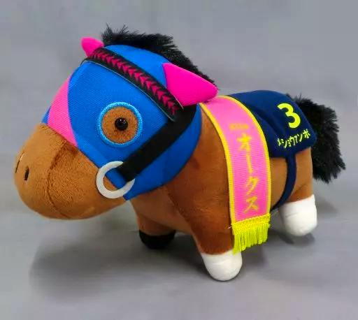 Plush toy of Meisho Manbo wearing blue-and-pink headgear, a pink sash with Japanese text, and a navy saddlecloth numbered 3.
