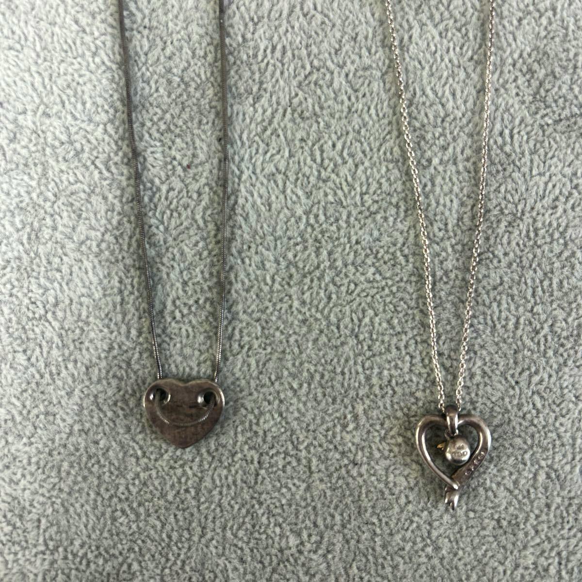 Two silver heart-motif necklaces (two-piece set) laid flat on a gray towel background, showing both pendants and chains.