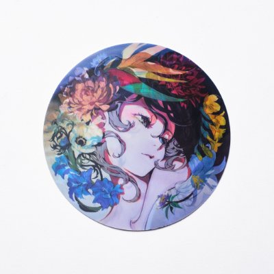 Round lenticular postcard showing a colorful illustrated woman's face with floral and feather motifs on a white background.