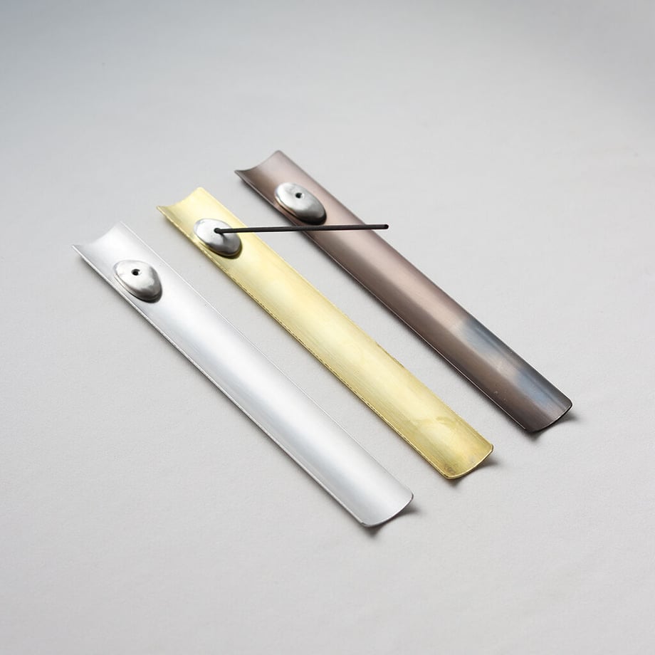 Three metal incense holders (tin, brass, and dark finish) arranged on a light background, one holding an incense stick on its small saucer.