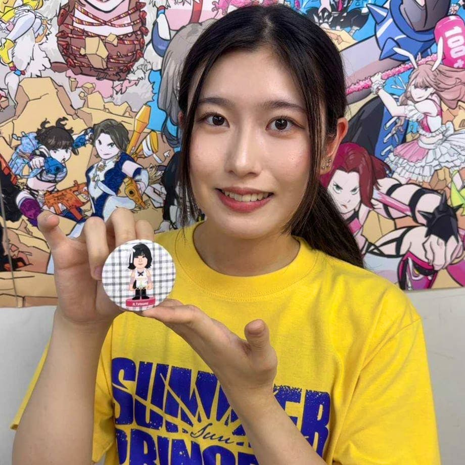 A person wearing a yellow shirt holds up a round TJPW illustration can badge showing a chibi wrestler on a checkered background.