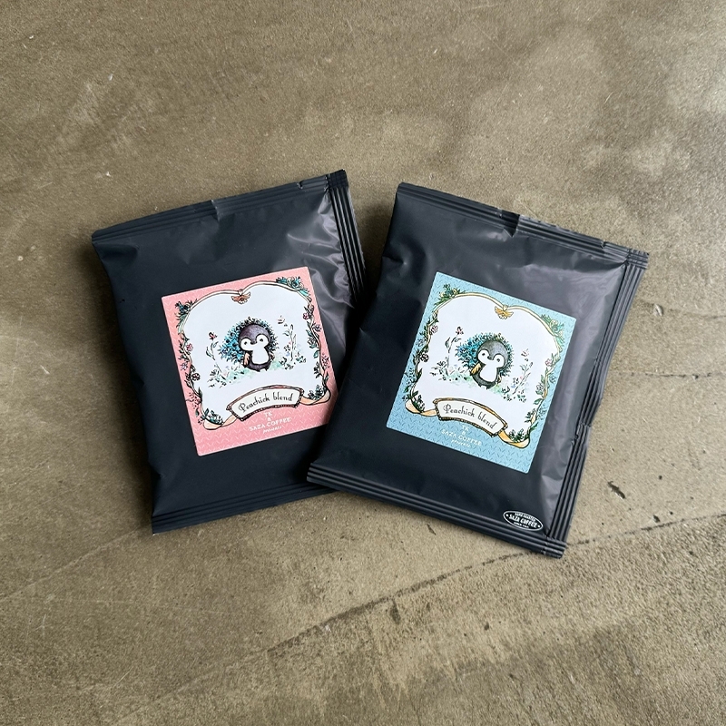 Two black packets of 'peachick Blend' instant coffee with illustrated labels showing small bird characters, placed on a textured surface.