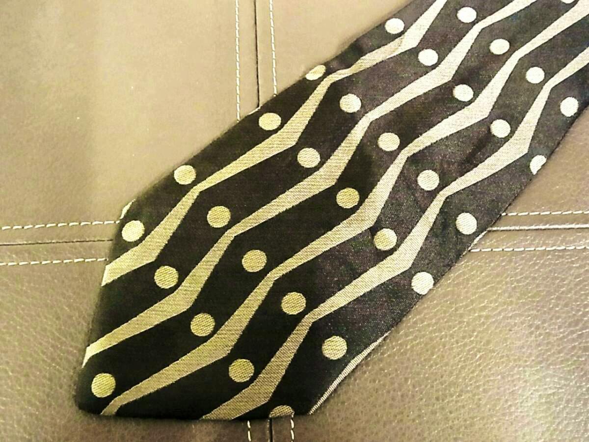 Close-up of a silk necktie tip showing a black background with gold wavy stripes and gold polka dots.