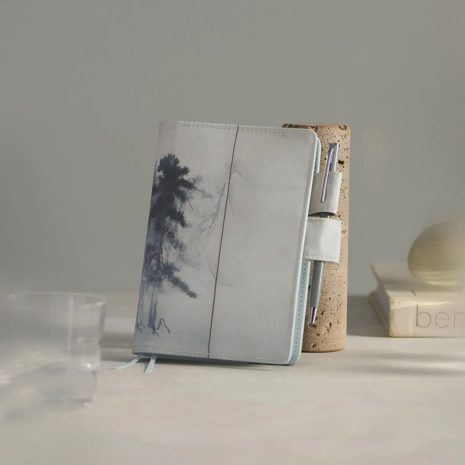 A closed A6 diary with the Hasegawa Tohaku Pine Forest cover, standing upright with a pen in its side loop and blurred props in the background.
