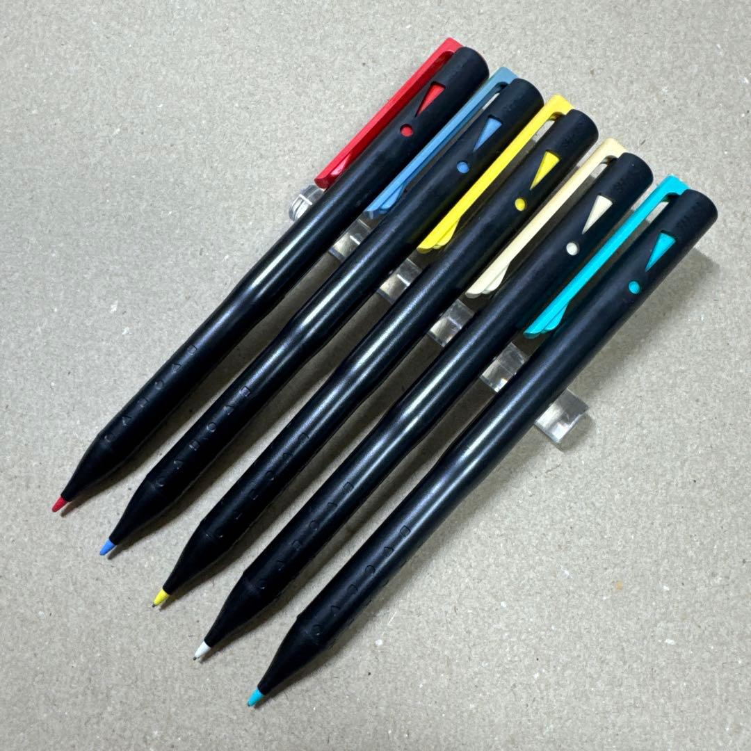 Five black mechanical pencils with different colored clips and colored tips arranged on a plain surface.