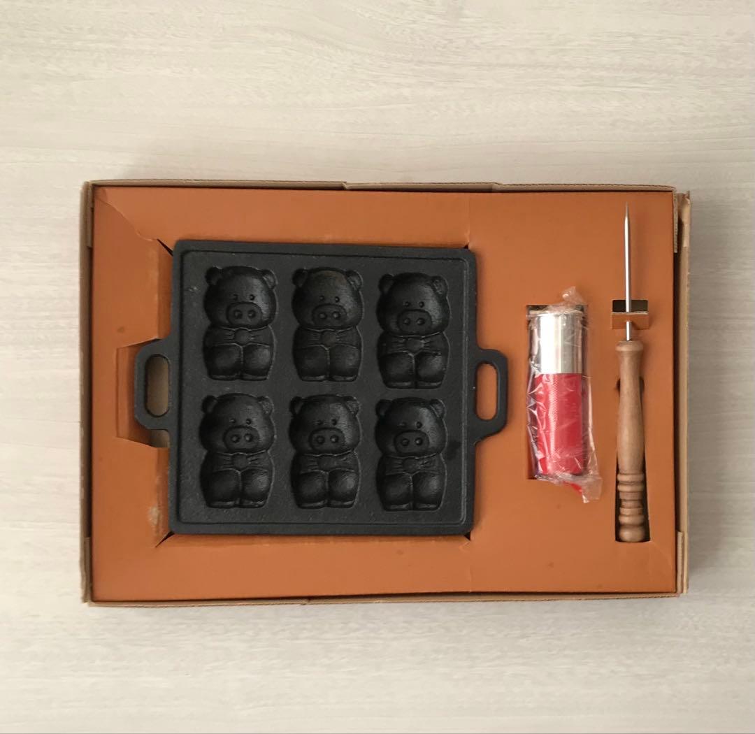 Top-down photo of the grill plate set showing a cast-iron mold plate with six doll-shaped cavities alongside a wooden handle and a wrapped accessory in its box.