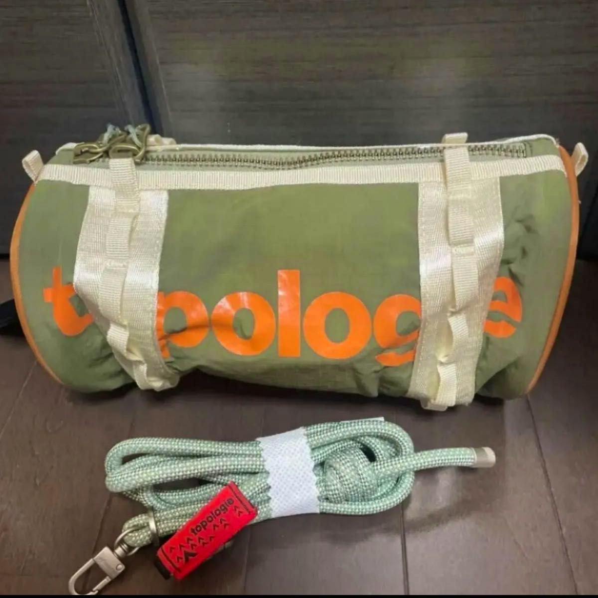 Olive-green cylindrical Topologie bag with orange logo, cream webbing straps, and a coiled green shoulder strap placed in front.