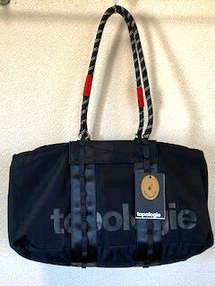 Front view of a black Topologie Summit small duffle bag with shoulder straps and a hanging product tag.