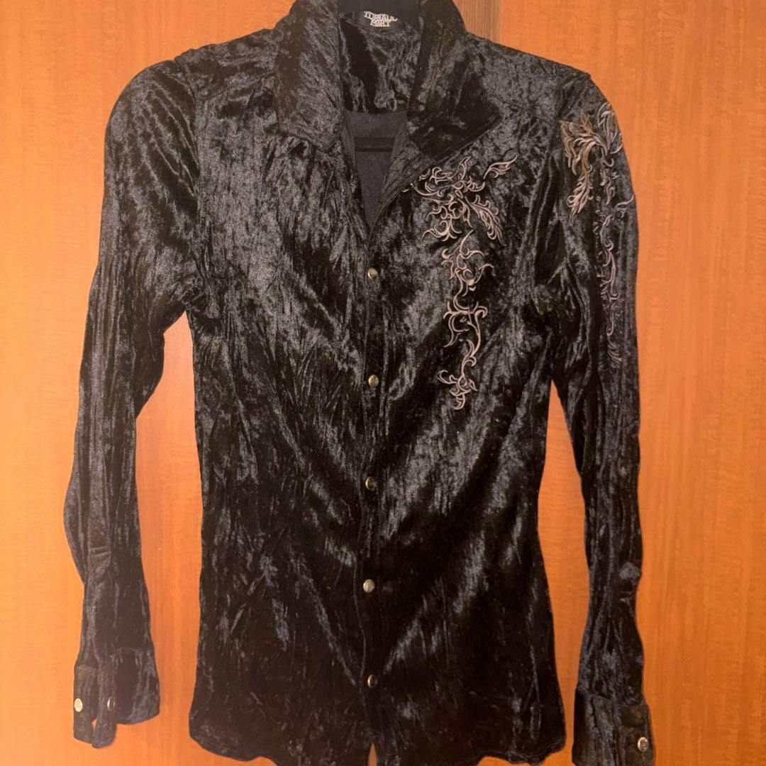 Front view of a black shiny open-collar men's shirt with embroidery and buttons hanging against a wooden background.