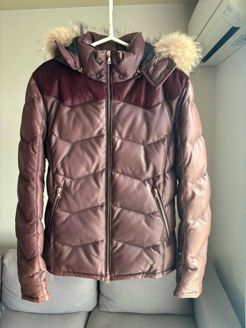 Front view of a brown lambskin leather down jacket with a fur-trimmed hood hanging on a hanger.