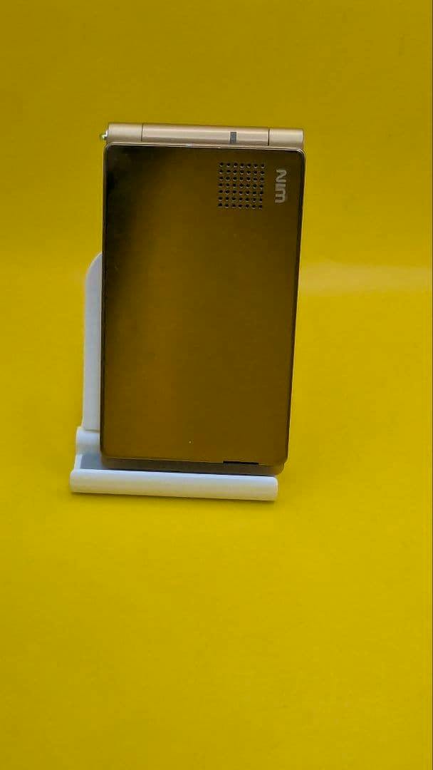 Closed TOSHIBA W55T au flip mobile phone showing the back on a white stand against a yellow background.