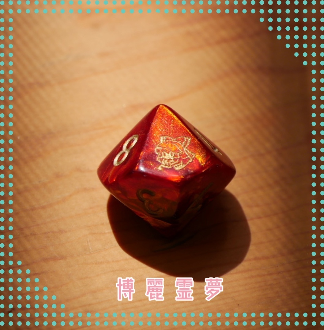 Close-up photo of a red 10-sided plastic die with gold numbers and a small printed Touhou Project character icon on a wooden surface.