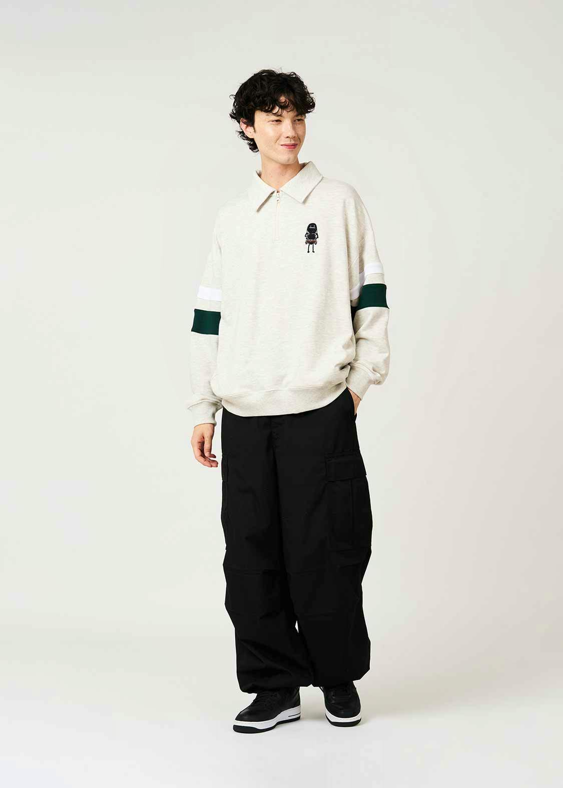 Front view of the 訓練兵シャドー ハーフジップスウェット worn by a model, showing a light-colored half-zip sweatshirt with collar, a small chest graphic, and green and white sleeve stripes.