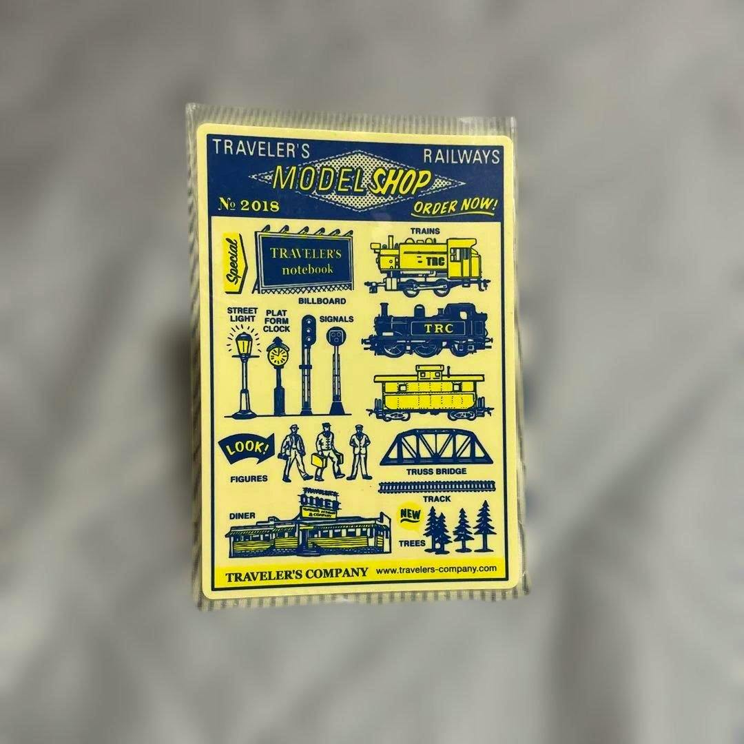 Front of the TRAVELER'S COMPANY 下敷き 2018 passport-size acrylic plate showing a yellow card with blue train-themed illustrations in its packaging.