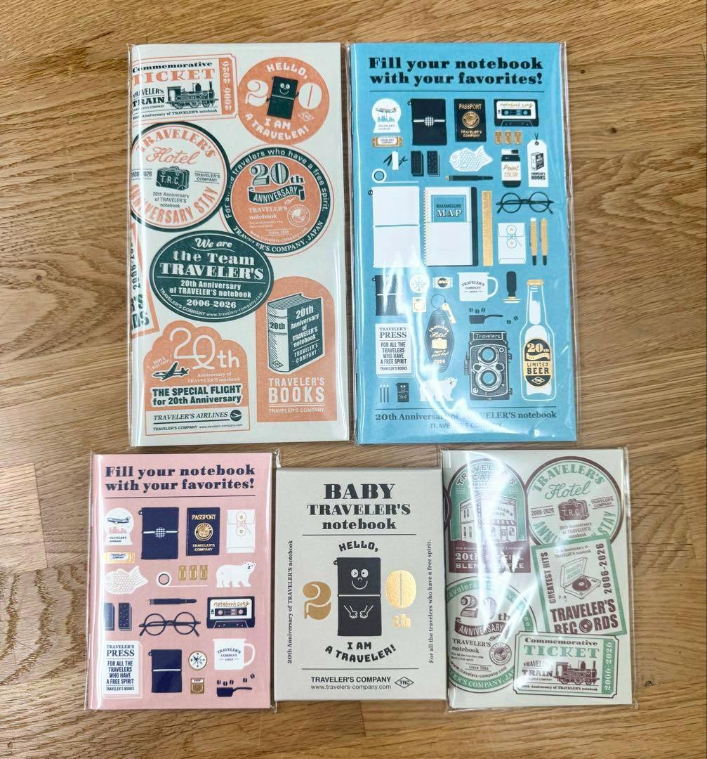 Five sealed Traveler's Notebook Baby 20th anniversary notebooks and decorative inserts arranged on a wooden floor.