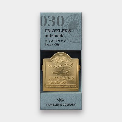 A brass Traveler's Notebook clip with an engraved airplane design shown in its retail box packaging.