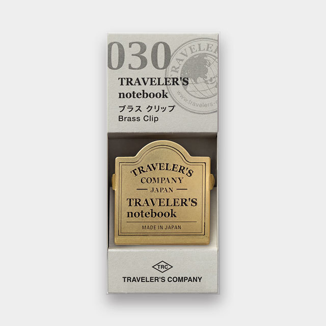 A brass Traveler's notebook clip in its cardboard packaging showing TRAVELER'S notebook and TRAVELER'S COMPANY branding.