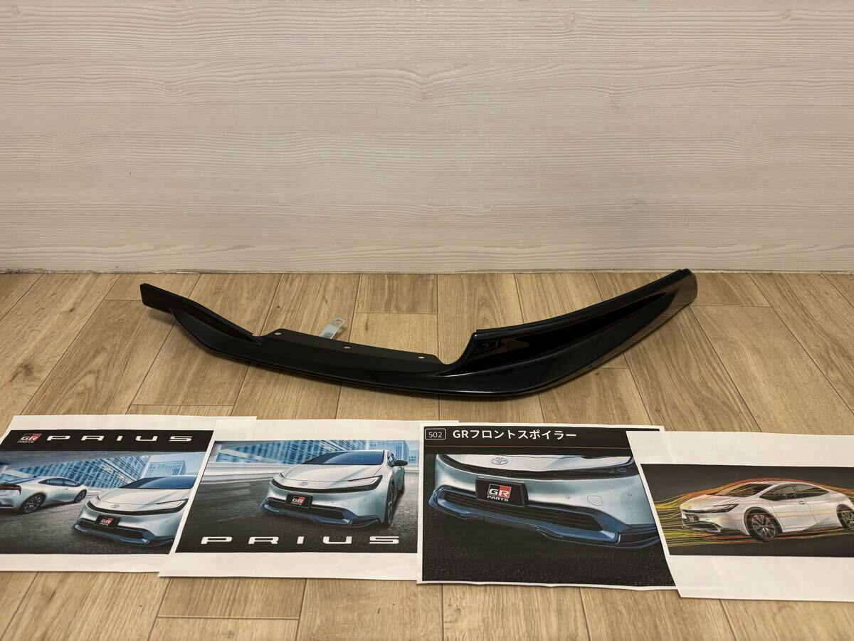 Black front lip spoiler for a Toyota Prius shown on a wood floor with printed Prius promotional images placed in front.