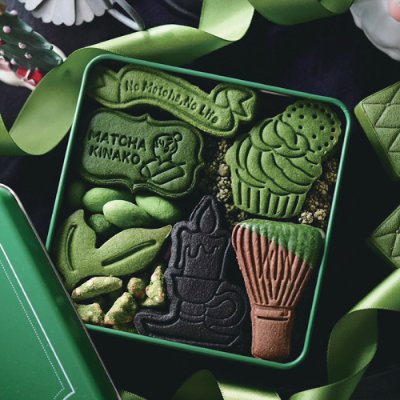 Open green tin showing assorted MATCHA KINAKO cookies shaped like a cupcake, tea whisk, tea bowl, and decorative motifs.
