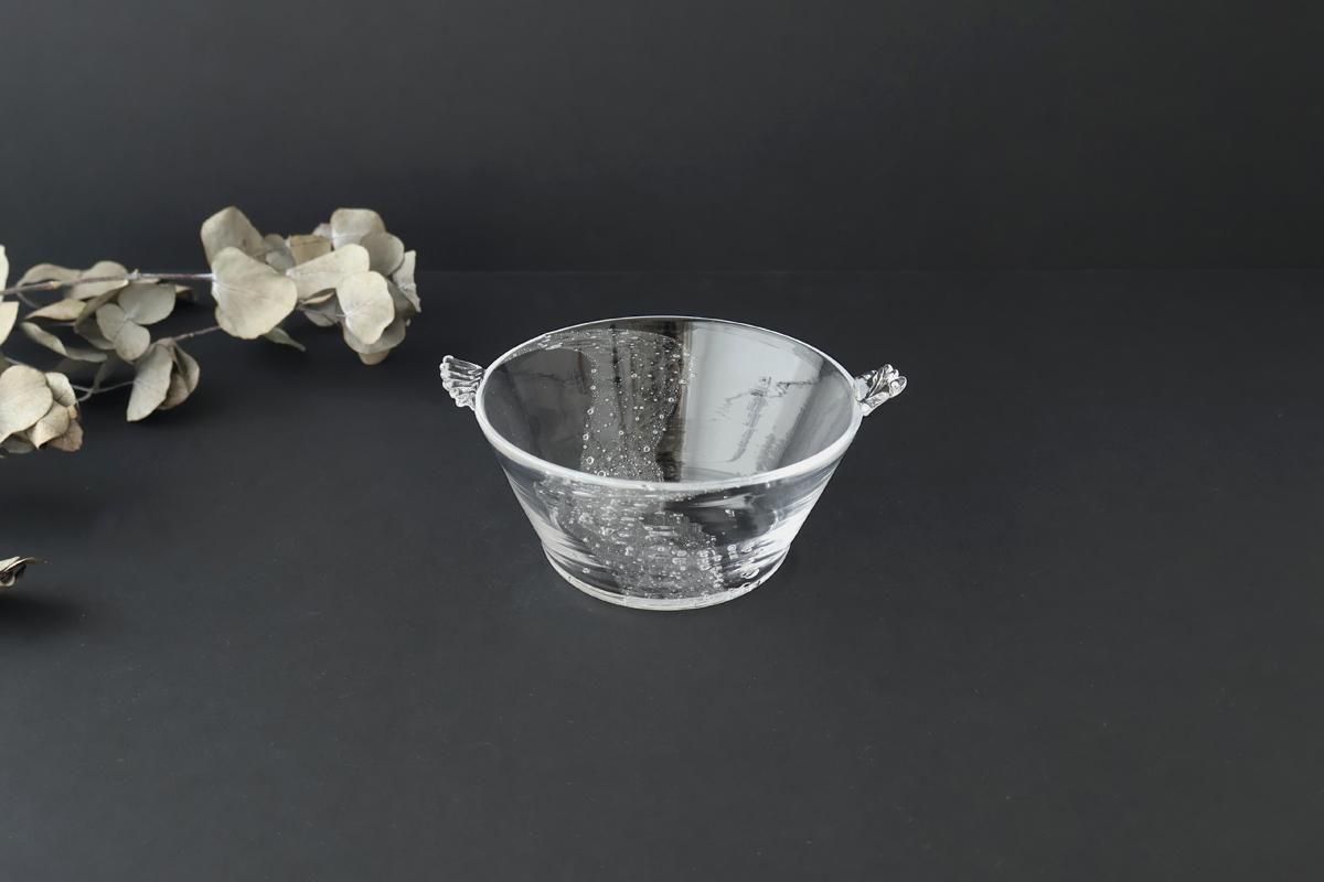 Clear glass soup bowl with small decorative handles on a dark background next to dried leaves.