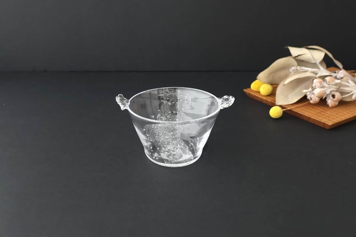 Clear small glass bowl with two decorative side handles on a black surface with dried flowers to the right.