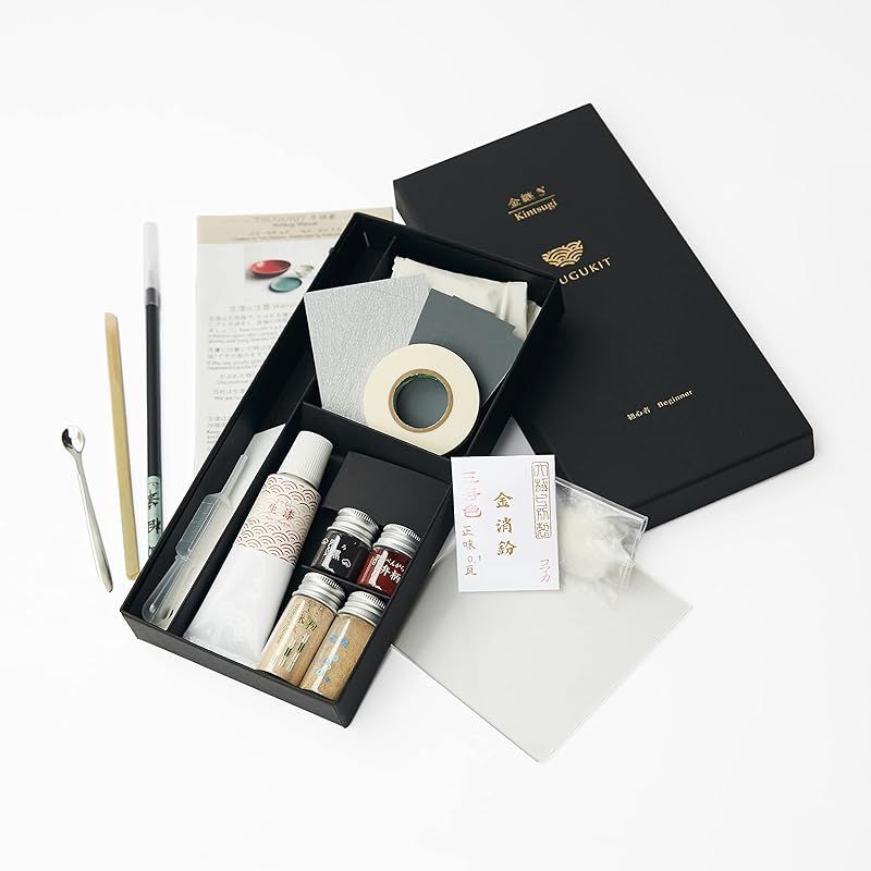 TSUGUKIT kintsugi (gold) repair kit arranged with a black box, bottles, tubes, tape, brushes, and tools.