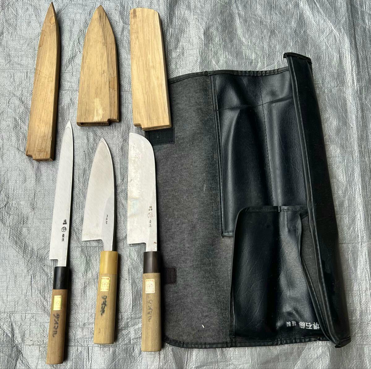 Three chef knives with wooden handles and wooden sheaths laid beside an open black knife roll case on a gray surface.