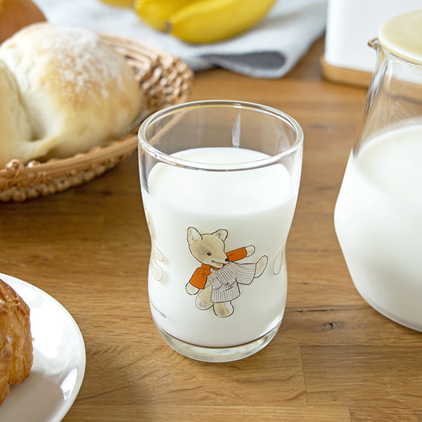A drinking glass with an illustrated character, filled with milk, on a wooden table beside bread and a milk jug.
