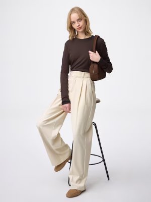 Female model wearing cream wide pleated women's pants and a brown long-sleeve top, posing with one foot on a stool.
