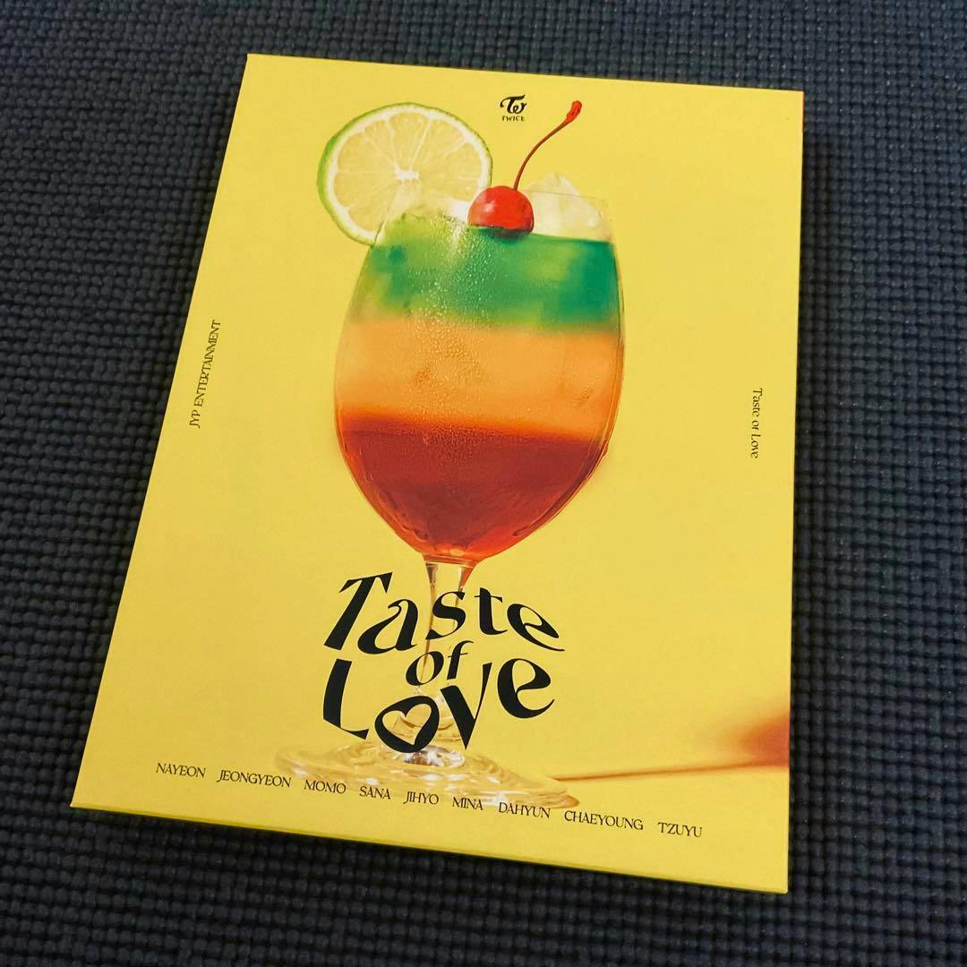 Front cover of the TWICE "Taste of Love" monograph showing a colorful layered cocktail illustration on a yellow background.